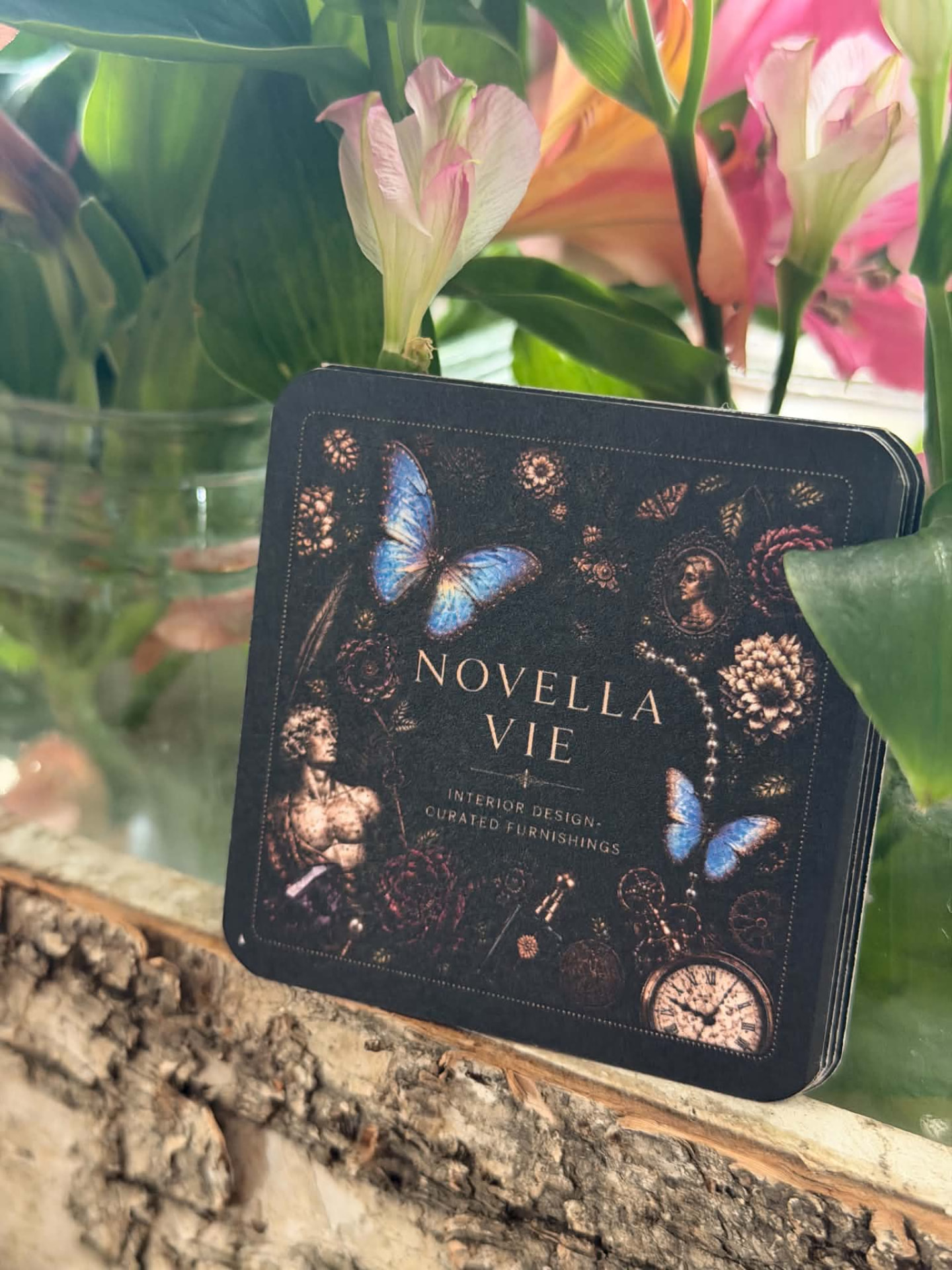 A stack of Novella Vie cards with gold botanical artwork and a blue butterfly, resting on driftwood beside fresh flowers