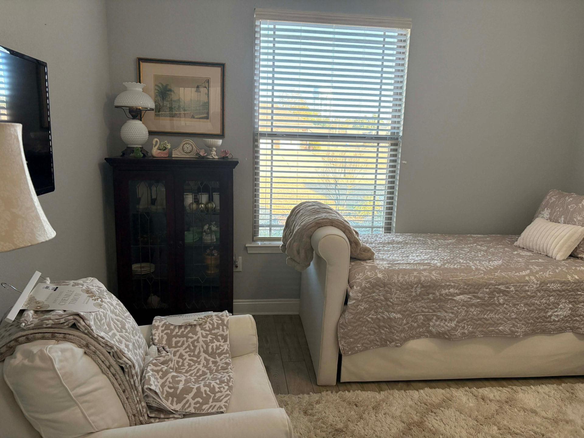 The bedroom before: a cream daybed, pale walls, and a dark glass-front cabinet beside the window